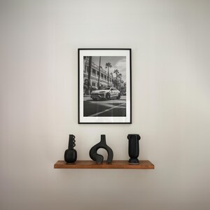 May include: A black and white photograph of a white SUV driving down a street lined with palm trees. The photo is framed in a black frame and is hanging on a white wall above a wooden shelf with three black vases.