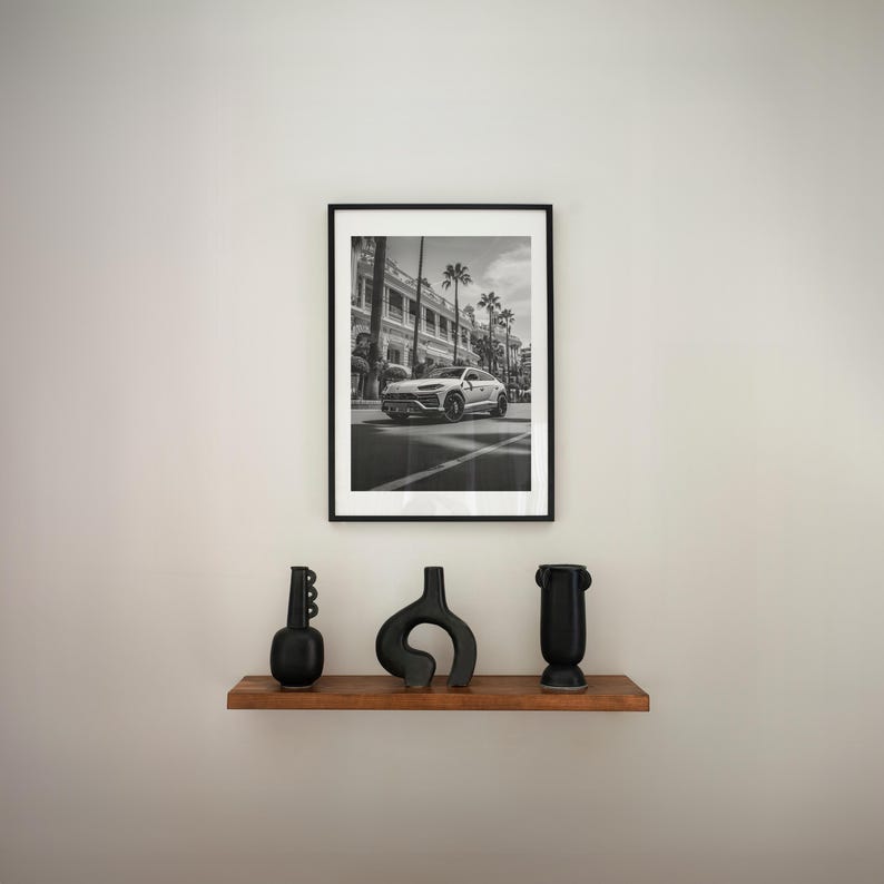 May include: A black and white photograph of a white SUV driving down a street lined with palm trees. The photo is framed in a black frame and is hanging on a white wall above a wooden shelf with three black vases.