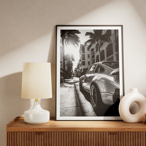 May include: Black and white photograph of a classic sports car parked on a street lined with palm trees and buildings. The car is facing away from the camera, and the sun is shining brightly in the background.