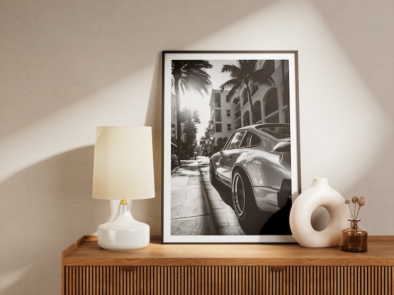 May include: Black and white photograph of a classic sports car parked on a street lined with palm trees and buildings. The car is facing away from the camera, and the sun is shining brightly in the background.