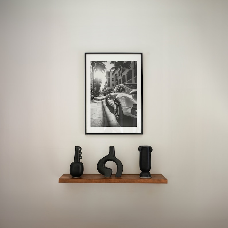 May include: A black and white photograph of a silver car driving down a palm tree lined street. The photo is framed in a black frame and is hanging on a white wall above a wooden shelf with three black vases.