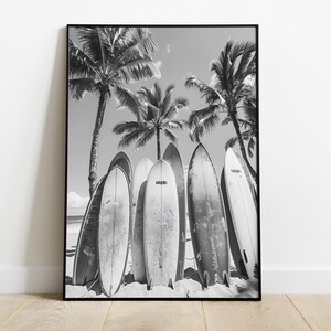 May include: Black and white photograph of five surfboards leaning against palm trees on a beach.