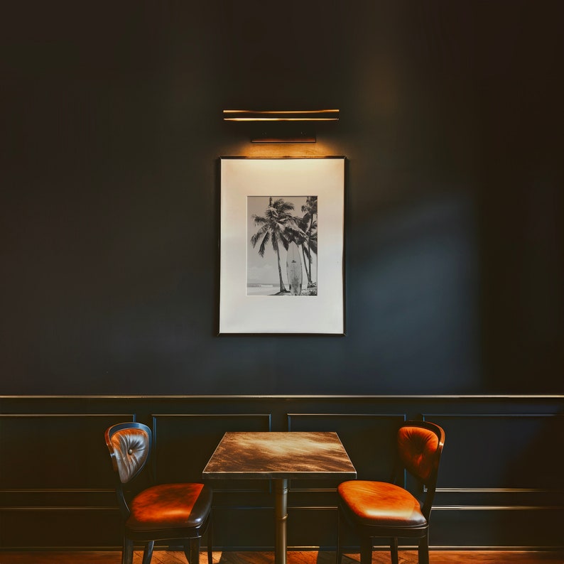 May include: A black and white photograph of palm trees and a surfer on a beach hangs on a dark wall above a small table with two brown leather chairs.