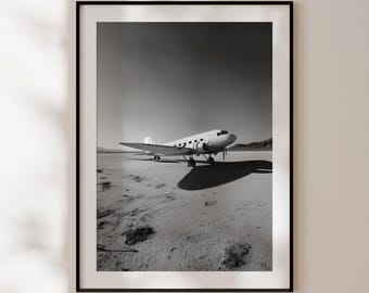 Op de afbeelding: Zwart-wit ingelijste print van een vintage vliegtuig op een droge zoutvlakte. Het vliegtuig, een klassiek propellermodel, werpt een lange schaduw. De afbeelding toont een contrast tussen de witte romp van het vliegtuig en de donkere achtergrond.