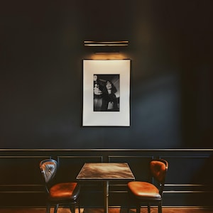 May include: A framed black and white photograph hangs on a dark wall, illuminated by a brass light fixture. Below, a small square table with a patterned top and two leather chairs with tufted backs complete the scene.