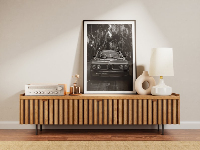Puede incluir: Una fotograf&iacute;a en blanco y negro de un coche cl&aacute;sico aparcado en una zona arbolada. La foto est&aacute; enmarcada y expuesta en una estanter&iacute;a de madera con un sistema est&eacute;reo y una l&aacute;mpara.