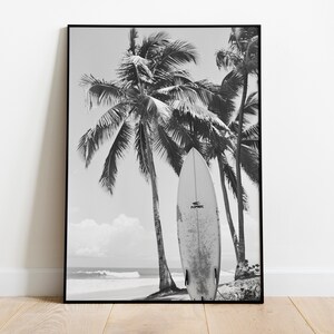 May include: Black and white photograph of a surfboard leaning against palm trees on a beach. The surfboard is white with black text that reads "SURFSKATE".