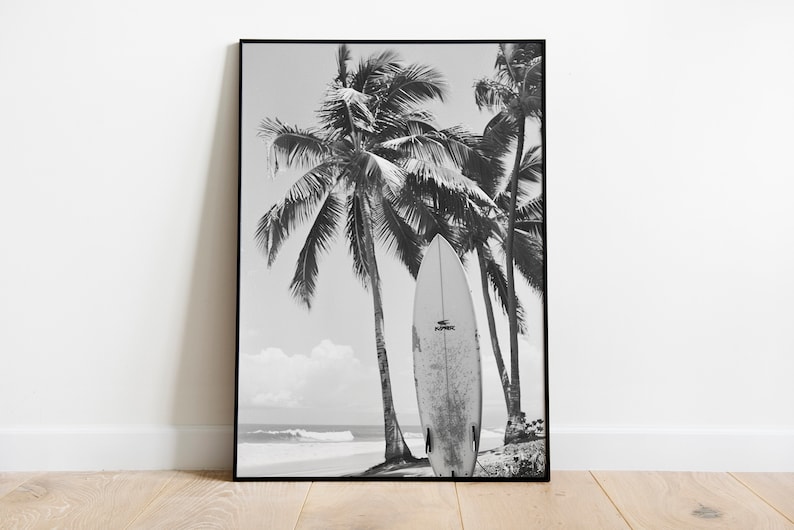 May include: Black and white photograph of a surfboard leaning against palm trees on a beach. The surfboard is white with black text that reads "SURFSKATE".