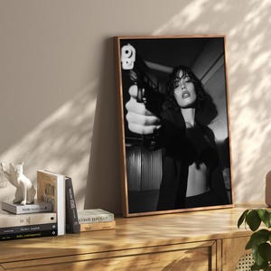 May include: Black and white framed print featuring a person holding a handgun, with a focus on the weapon. The image is displayed on a wooden cabinet, alongside books and decorative items.