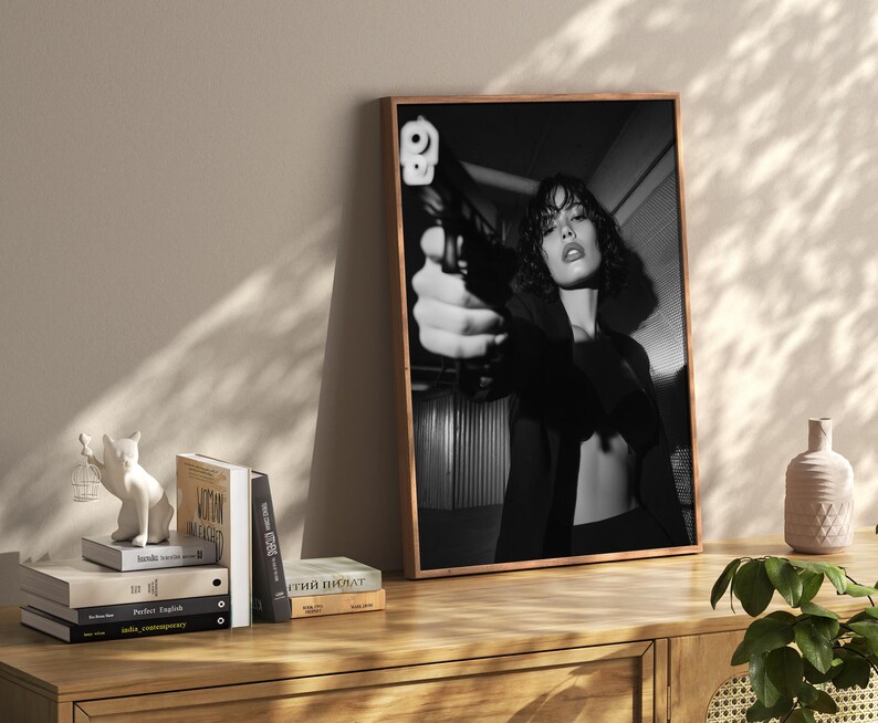 May include: Black and white framed print featuring a person holding a handgun, with a focus on the weapon. The image is displayed on a wooden cabinet, alongside books and decorative items.