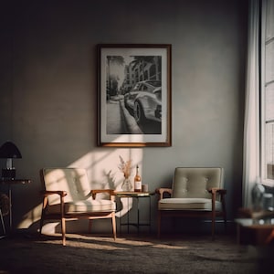 May include: Two beige armchairs with wooden frames and a small black metal side table with a bottle of wine and a vase of dried flowers in a living room with a framed black and white photograph of a car on the wall.