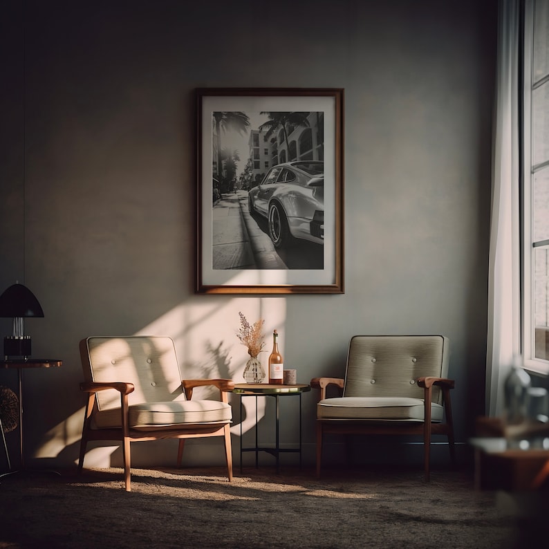 May include: Two beige armchairs with wooden frames and a small black metal side table with a bottle of wine and a vase of dried flowers in a living room with a framed black and white photograph of a car on the wall.