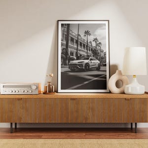 May include: A wooden media console with a black stereo receiver and a black and white framed photograph of a white car driving down a street with palm trees. A white ceramic lamp with a white shade sits on the console.