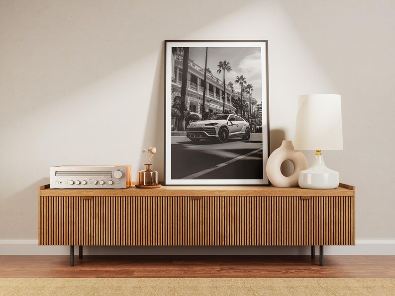May include: A wooden media console with a black stereo receiver and a black and white framed photograph of a white car driving down a street with palm trees. A white ceramic lamp with a white shade sits on the console.