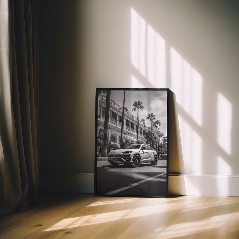 May include: A black and white photograph of a white SUV driving down a street lined with palm trees and buildings. The photo is framed in a black frame and is hanging on a white wall.