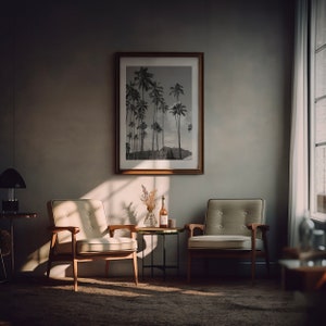 May include: Two mid-century modern chairs with light beige upholstery and wooden frames sit in a room with a black and white photograph of palm trees hanging on the wall. A small black metal side table with a bottle of wine and a vase of dried flowers sits between the chairs.