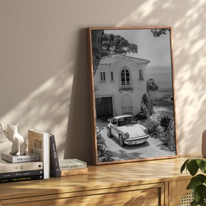 K&ouml;nnte beinhalten: Schwarzwei&szlig;foto eines wei&szlig;en Oldtimers, der vor einem wei&szlig;en Haus mit einem Steinbogen geparkt ist. Das Haus befindet sich auf einer Klippe mit Blick auf das Meer.