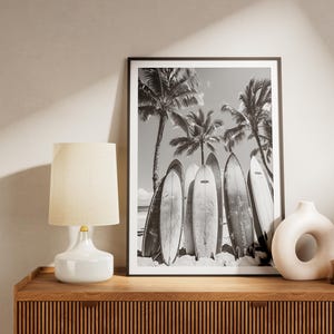 May include: Black and white photograph of surfboards leaning against palm trees on a beach. The photo is framed in a black frame and is displayed on a wooden shelf with a white lamp and a white ceramic vase.