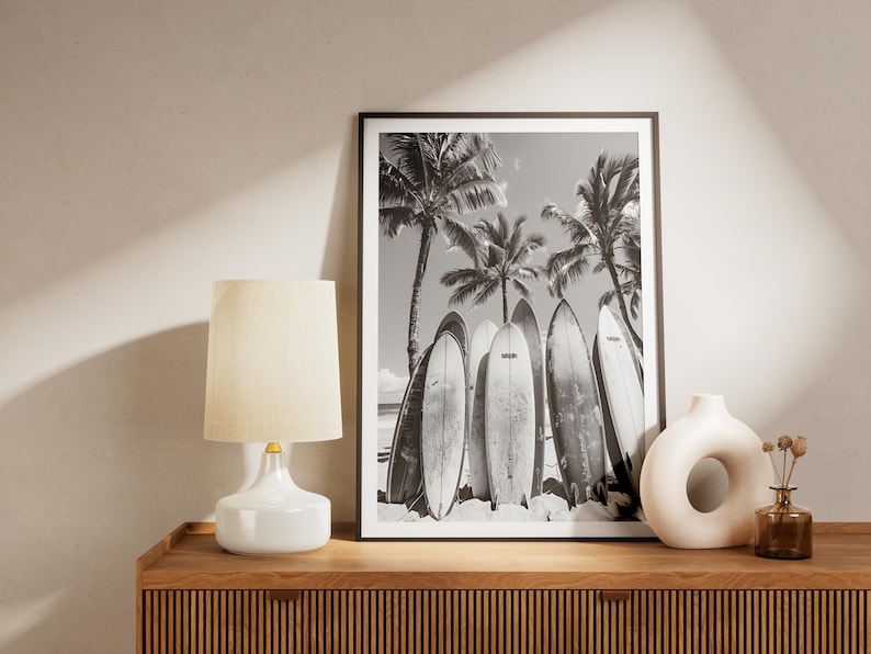 May include: Black and white photograph of surfboards leaning against palm trees on a beach. The photo is framed in a black frame and is displayed on a wooden shelf with a white lamp and a white ceramic vase.