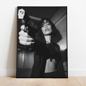 May include: Black and white framed print of a person holding a handgun, aiming it directly at the viewer. The person is wearing a dark blazer. The image is set against a textured background, creating a dramatic and intense visual.