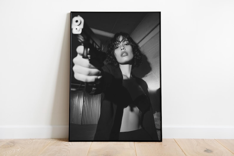 May include: Black and white framed print of a person holding a handgun, aiming it directly at the viewer. The person is wearing a dark blazer. The image is set against a textured background, creating a dramatic and intense visual.
