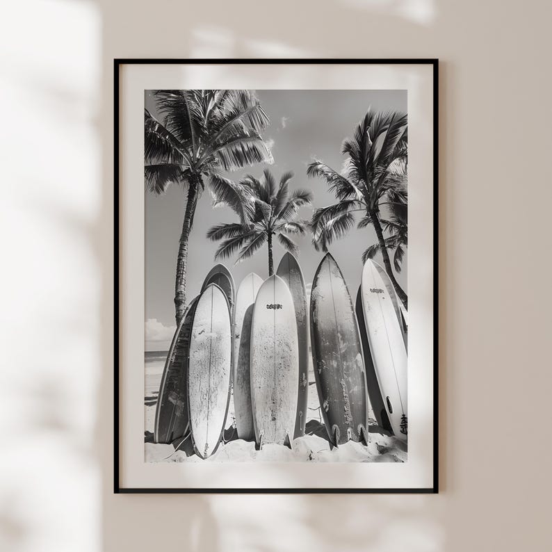 May include: Black and white photograph of six surfboards leaning against each other on a sandy beach. Palm trees are in the background.