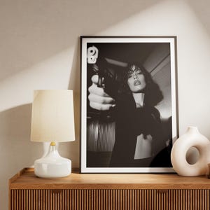 May include: Black and white framed print of a woman holding a gun. A white lamp with a beige shade, a white vase, and a brown vase with dried flowers sit on a wooden cabinet. The cabinet has a ribbed design.
