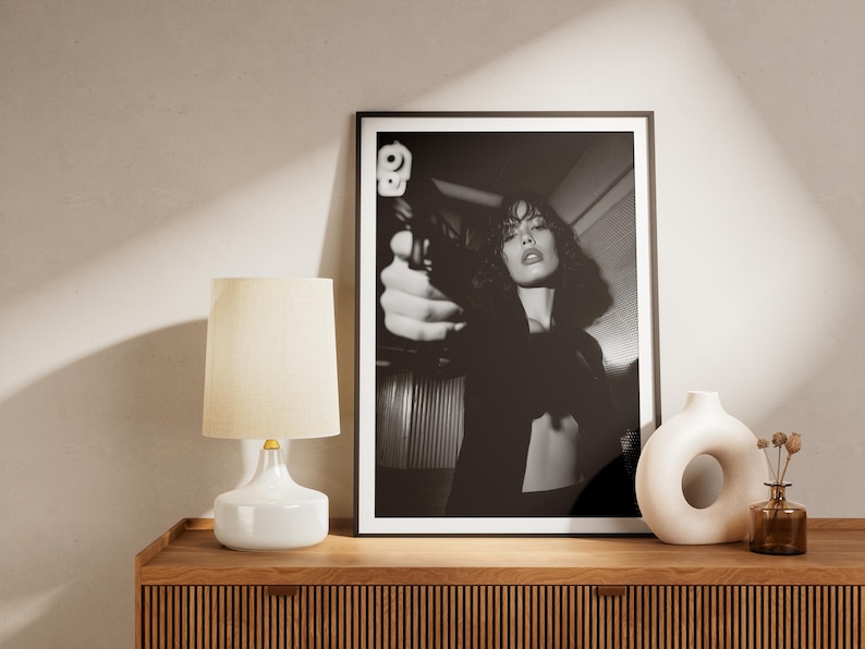 May include: Black and white framed print of a woman holding a gun. A white lamp with a beige shade, a white vase, and a brown vase with dried flowers sit on a wooden cabinet. The cabinet has a ribbed design.