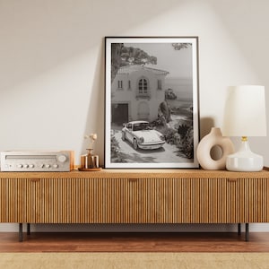 K&ouml;nnte beinhalten: Ein Schwarzwei&szlig;foto eines wei&szlig;en Autos, das vor einem Haus mit Blick auf das Meer geparkt ist. Das Foto ist gerahmt und auf einem Holzregal mit einer Lampe und einer Vase mit Blumen ausgestellt.