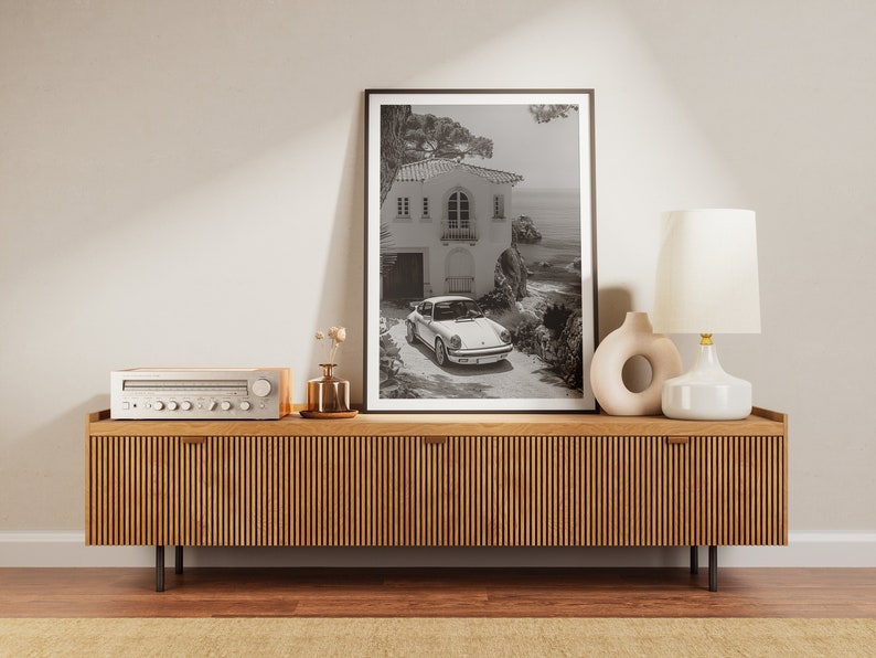 K&ouml;nnte beinhalten: Ein Schwarzwei&szlig;foto eines wei&szlig;en Autos, das vor einem Haus mit Blick auf das Meer geparkt ist. Das Foto ist gerahmt und auf einem Holzregal mit einer Lampe und einer Vase mit Blumen ausgestellt.