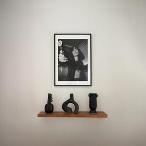 May include: A black and white framed photograph of a person holding a gun. Below the photograph, a wooden shelf displays three black decorative vases of varying shapes. The vases add a touch of modern art to the space.