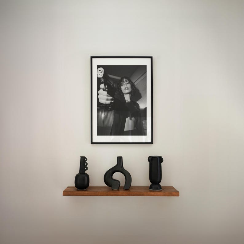 May include: A black and white framed photograph of a person holding a gun. Below the photograph, a wooden shelf displays three black decorative vases of varying shapes. The vases add a touch of modern art to the space.