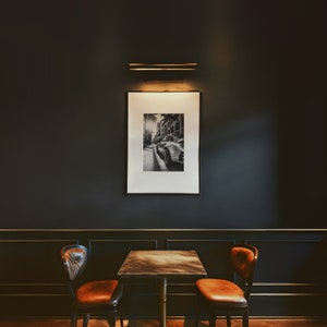 May include: A black and white photograph of a car driving down a street is framed and hanging on a dark wall. Two brown leather chairs with a dark wood table are in front of the picture.