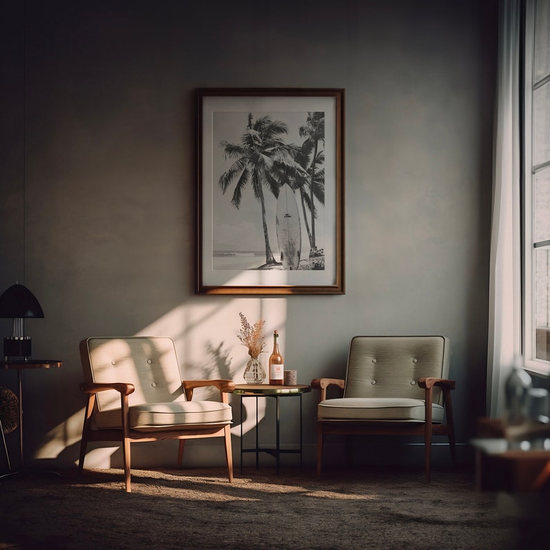 May include: Two mid-century modern armchairs with light beige upholstery and wooden frames, a small black metal side table with a bottle of wine and a vase of dried flowers, and a framed black and white photograph of palm trees and a surfboard on a beach.