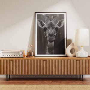 May include: A black and white photograph of a giraffe's head with its tongue sticking out, framed and displayed on a wooden media console with a vintage stereo receiver, a small vase with dried flowers, and a white ceramic lamp with a white lampshade.
