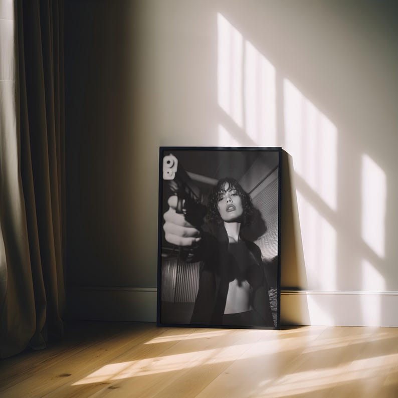 May include: Black and white framed print of a woman holding a gun. The image is leaning against a white wall on a wooden floor. The woman is wearing a black jacket and has curly hair. The image is lit by natural light.