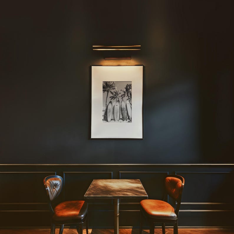 May include: A black and white photograph of palm trees and surfboards hangs on a dark wall above a wooden table with two brown leather chairs. The table has a dark wood top and a metal base.