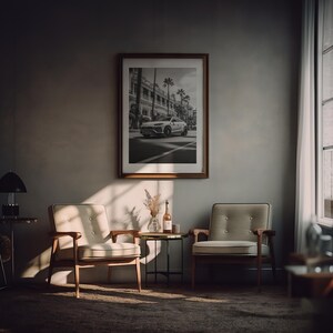 May include: A black and white photograph of a car driving down a street in a city. The photo is framed in a brown wood frame and hanging on a wall in a living room. Two beige armchairs are positioned in front of the photo, and a small table with a bottle of wine and a vase of flowers sits between them.