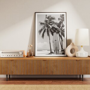 May include: A wooden media console with a black and white photograph of palm trees and a surfboard on the beach. The console has a silver stereo receiver and a white ceramic lamp with a gold base.