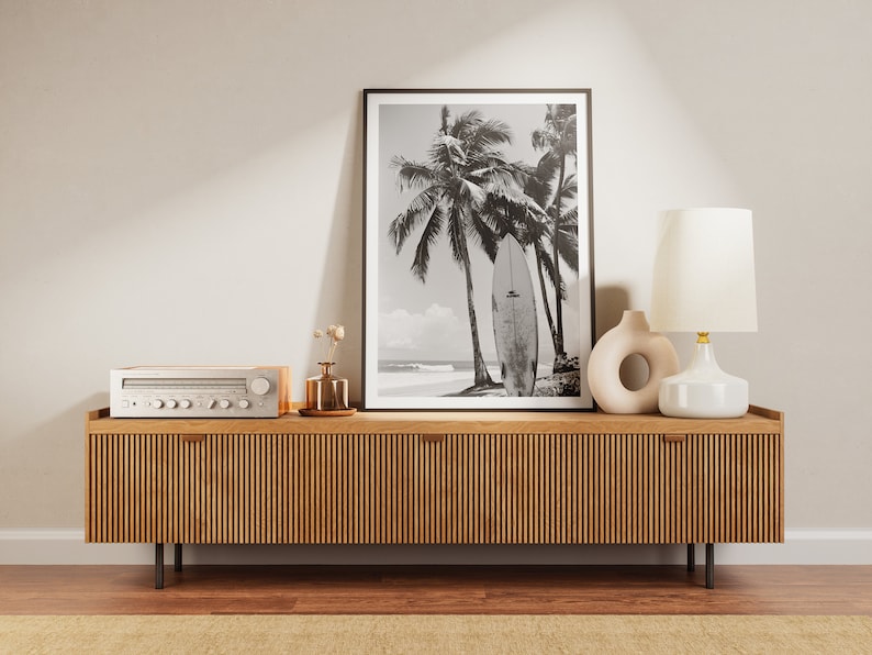 May include: A wooden media console with a black and white photograph of palm trees and a surfboard on the beach. The console has a silver stereo receiver and a white ceramic lamp with a gold base.