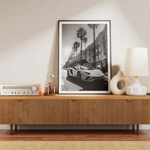 May include: A black and white photograph of a white sports car driving down a street lined with palm trees and buildings. The photo is framed and hanging on a wall above a wooden entertainment unit with a stereo receiver and two white ceramic vases.