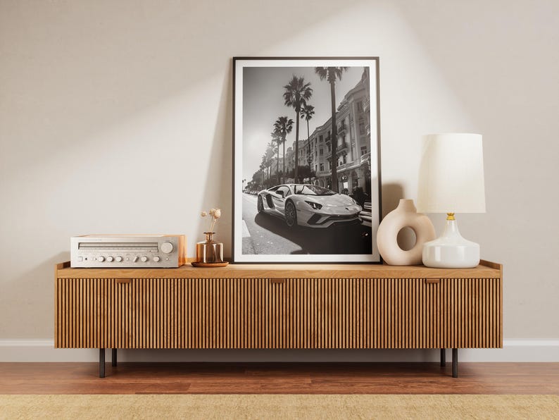 May include: A black and white photograph of a white sports car driving down a street lined with palm trees and buildings. The photo is framed and hanging on a wall above a wooden entertainment unit with a stereo receiver and two white ceramic vases.