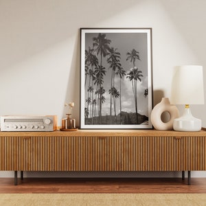 May include: A wooden media console with a light wood finish and vertical grooves. The console has two drawers and is decorated with a black and white framed print of palm trees, a white ceramic lamp with a gold base, and a white ceramic vase with a circular cutout.
