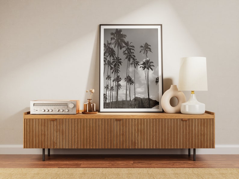 May include: A wooden media console with a light wood finish and vertical grooves. The console has two drawers and is decorated with a black and white framed print of palm trees, a white ceramic lamp with a gold base, and a white ceramic vase with a circular cutout.