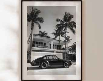 May include: A black and white photograph of a classic car parked in front of a white house with palm trees in the background.
