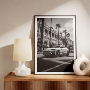 May include: Black and white photograph of a white SUV driving down a street lined with palm trees and buildings. The photo is framed in a black frame and is hanging on a wall above a wooden dresser.