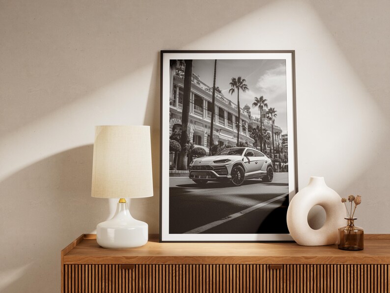 May include: Black and white photograph of a white SUV driving down a street lined with palm trees and buildings. The photo is framed in a black frame and is hanging on a wall above a wooden dresser.