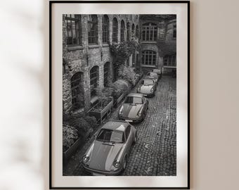 May include: A black and white photograph of five vintage Porsche cars parked in a row in a brick courtyard. The cars are all facing the same direction and are lined up in a straight line. The courtyard is surrounded by brick buildings with windows and a green ivy growing on the walls.