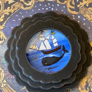 K&ouml;nnte beinhalten: Ein schwarzer Holzrahmen mit einem gewellten Rand umgibt eine gemalte Szene eines Schiffes, das auf einem blauen Meer segelt, unter dem ein Wal schwimmt. Das Gem&auml;lde ist mit "Jane" signiert.