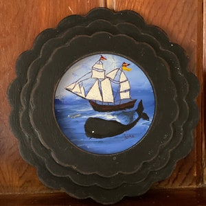 K&ouml;nnte beinhalten: Ein rundes Wandbild mit schwarzem Rahmen, das eine gemalte Szene eines Segelboots mit wei&szlig;en Segeln und einem Wal im blauen Ozean zeigt. Die Unterschrift des K&uuml;nstlers ist in der rechten unteren Ecke sichtbar.